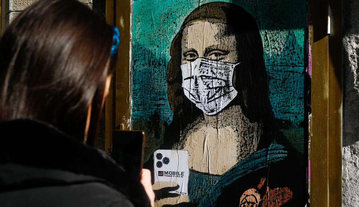 Seorang perempuan mengambil foto poster seniman Italia Salvatore Benintende yang menggambarkan Mona Lisa karya Leonardo da Vinci mengenakan masker dan memegang smartphone yang bertuliskan "Mobile World Virus" di sebuah jalan di kota Barcelona, Spanyol pada Selasa (18/2/2020). (PAU BARRENA/AFP)