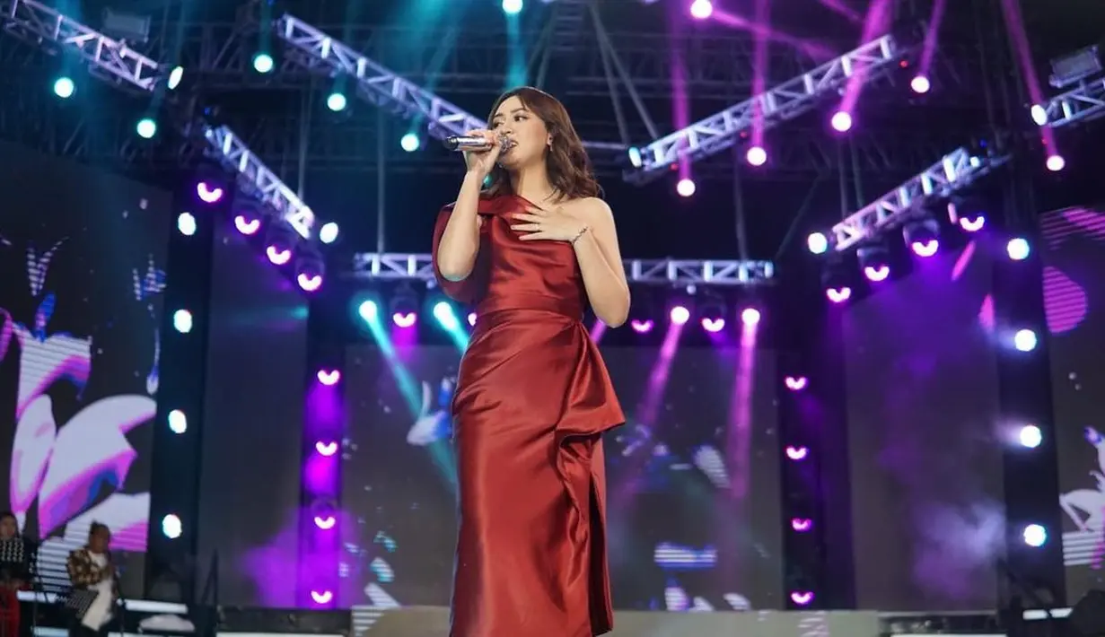 Happy Asmara tampil cantik di atas panggung mengenakan gaun merah. Gaun yang dikenakannya memiliki detail bahu asimetris dengan lipit di bagian pinggangnya. [Foto: Instagram/happy_asmara77]