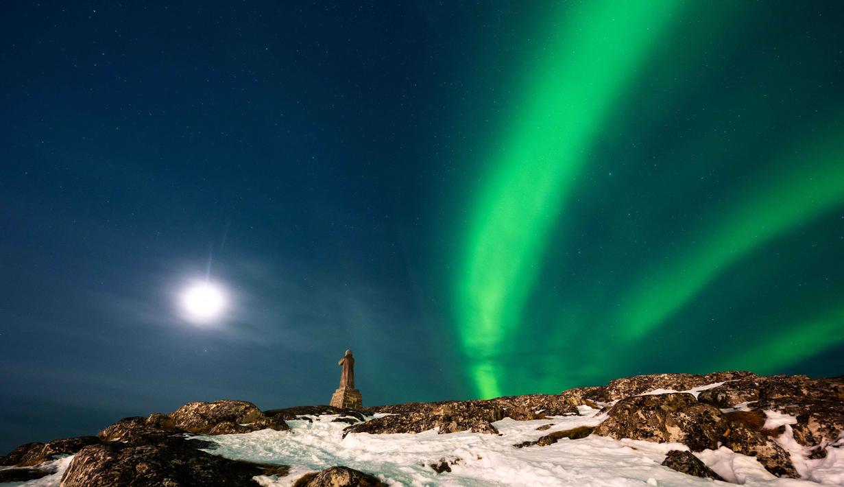 Fenomena ini terjadi ketika partikel bermuatan dari Matahari bertabrakan dengan medan magnet dan atmosfer bumi. Tampak dalam foto, Aurora Borealis menerangi langit malam di atas patung Hans Egede yang menghadap Nuuk, Greenland, pada 23 Januari 2026. (Jonathan NACKSTRAND/AFP)