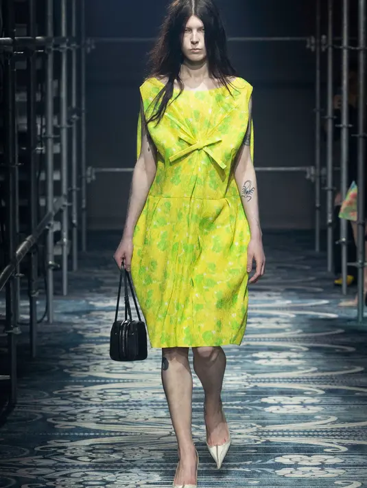 Pertunjukan ini digelar di ruang Deposito milik Fondazione Prada, di mana atmosfer raw dipadukan dengan elemen dekorasi yang halus. Struktur logam membentuk konfigurasi baru, sementara karpet karya Catherine Martin menambah sentuhan artistik pada ruangan. Kontras antara kekasaran dan kehalusan ini merefleksikan kompleksitas makna yang tertanam dalam setiap busana. [Dok/Prada].