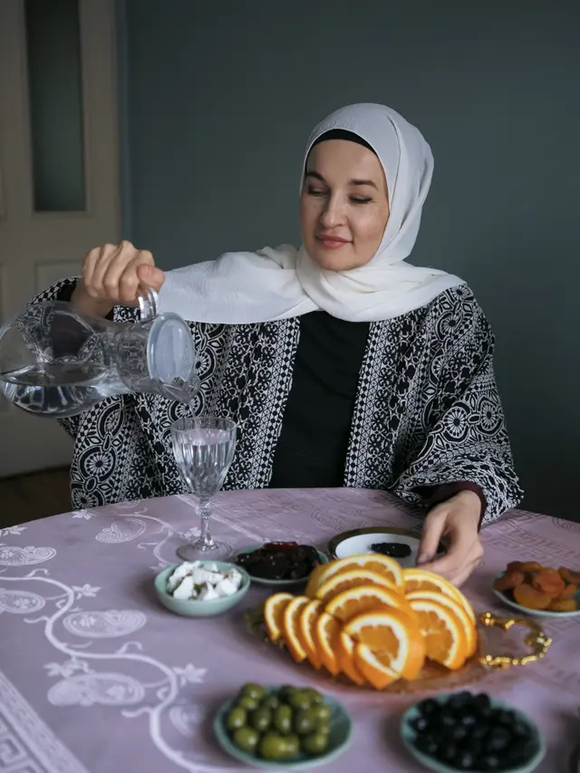 Seorang perempuan berhijab sedang duduk di meja makan dan menuangkan air ke gelas (Foto Dok: Freepik/freepik)