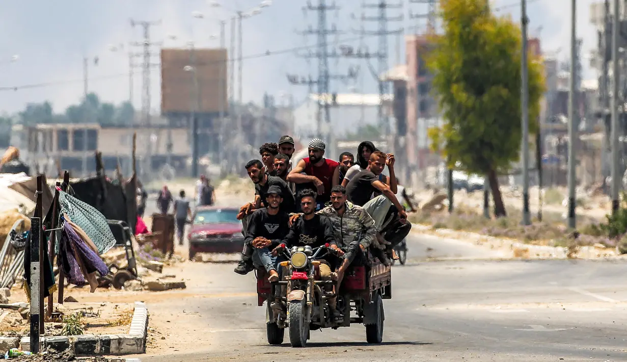 Para pria berkendara bersama di belakang gerobak roda tiga di sepanjang jalan Salaheddin, Deir el-Balah, Jalur Gaza tengah pada 21 Juli 2025, sementara di belakangnya mengepul asap akibat pemboman Israel. (Eyad BABA/AFP)