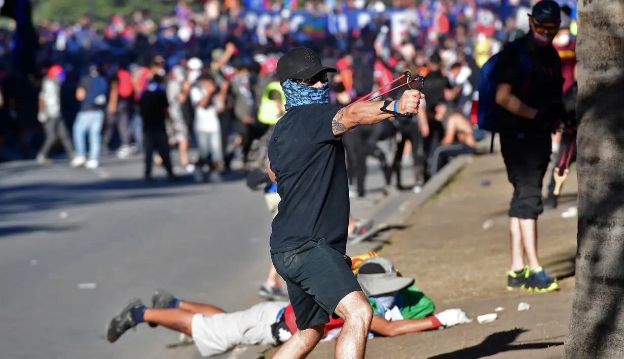Seorang demonstran menggunakan ketapel menyerang polisi anti huru hara selama protes terhadap kebijakan ekonomi pemerintah di Santiago (6/11/2019). Presiden Chile mengatakan "tidak menyembunyikan apa-apa" mengenai tuduhan bahwa polisi membunuh, menyiksa warga sipil. (AFP Photo/Martin Bernetti)