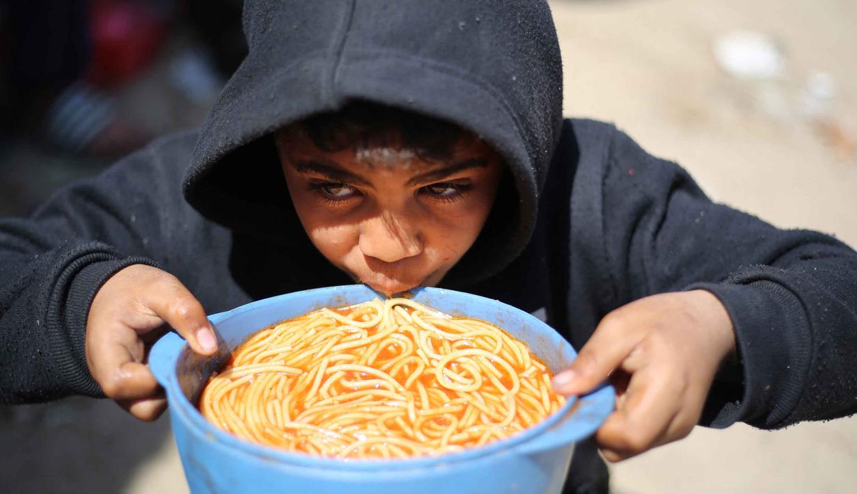 Patut dicatat, terdapat sekitar 1,9 juta dari total populasi 2,4 juta warga Gaza yang bertahan hidup di lokasi-lokasi pengungsian, banyak di antaranya tinggal di tenda darurat setelah rumah mereka hancur akibat konflik berkepanjangan antara Israel dan Hamas. Tampak dalam foto, seorang anak laki-laki Palestina yang mengungsi makan dari mangkuk setelah menerima makanan dari dapur amal di kamp pengungsi Nuseirat, Jalur Gaza Tengah pada 13 April 2026. (Eyad Baba/AFP)