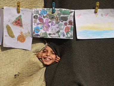 Seorang anak melihat melalui lubang di selimut selama pameran yang menampilkan koleksi gambar oleh anak-anak dan perempuan di sebuah sekolah yang diubah menjadi tempat penampungan bagi pengungsi Palestina di kawasan Al-Rimal, Kota Gaza, pada 5 November 2025. (Omar AL-QATTAA/AFP)