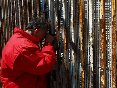 Seorang warga Meksiko terbujur kaku sambil memegang pagar perbatasan sambil berbincang dengan keluarganya di Playas Tijuana, Meksiko, (12/4). Tembok ini memisahkan antara Meksiko dan Amerika Serikat. (REUTERS / Jorge Duenes)
