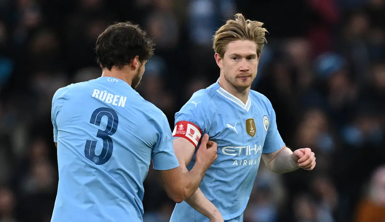 Pemain Manchester City, Ruben Dias (kiri), memberikan ban kapten kepada rekannya yakni Kevin De Bruyne saat memasuki lapangan pada menit ke-57 dalam pertandingan putaran ketiga Piala FA melawan Huddersfield Town yang berlangsung di Etihad Stadium, Manchester, Minggu (7/1/2024). (AFP/Paul Ellis)