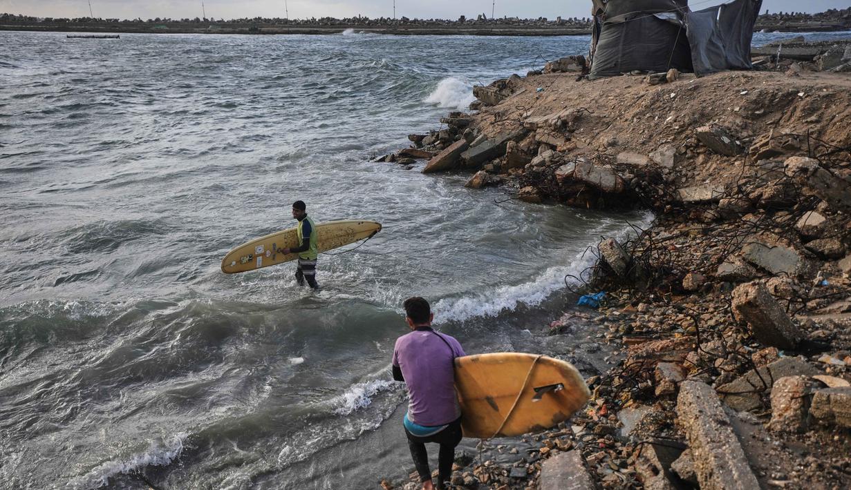 Di pantai Kota Gaza, beberapa warga Palestina menemukan kembali kenikmatan berselancar. Tampak dalam foto, warga Palestina, Khalil Abu Jayyab (depan) dan Tahseen Abu Assi membawa papan selancar mereka saat memasuki sepanjang garis pantai di Kota Gaza, Minggu, 28 Desember 2025. (AP Photo/Jehad Alshrafi)