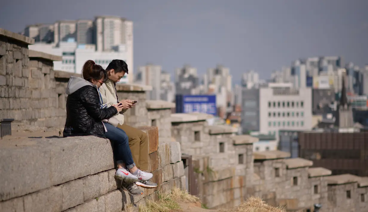 Suami istri melihat ponsel mereka ketika mereka duduk di lereng bukit yang menghadap ke cakrawala kota di Seoul, Korea Selatan (2/4). Korea Selatan sedang bersiap untuk meluncurkan jaringan seluler 5G pertama di dunia pada 5 April. (AFP Photo/Ed Jones)
