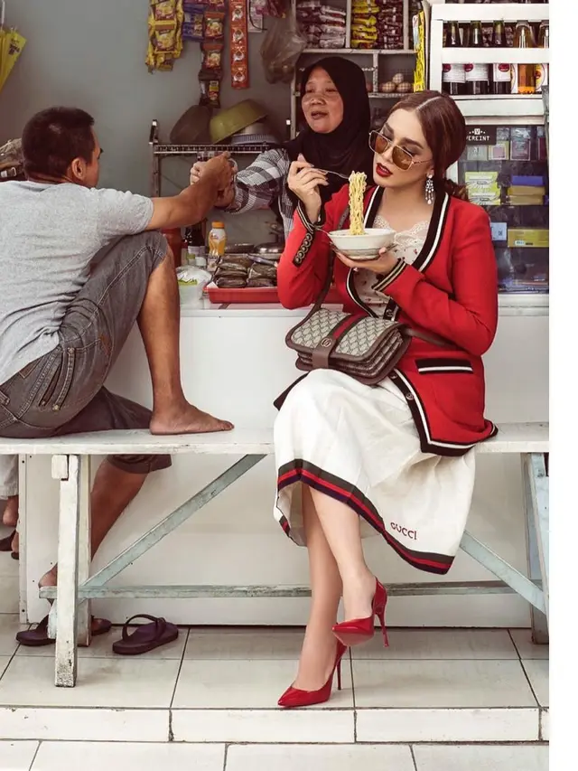 Ussy Sulistiawaty bergaya glamor sedang memegang mangkuk berisikan mi di dalam warung.