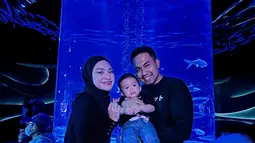 Entah disengaja atau tidak, Nathalie Holscher dan Fariz Utama kompak mengenakan celana jin biru yang dipadukan dengan atasan lengan panjang hitam tanpa kerah. Berfoto dengan latar aquarium vertikal plus tata cahaya lazuardi memberikan efek dramatis remang-remang. Menggendong baby Adzam, keduanya tersenyum lebar menikmati suasana. (Foto: Dok. Instagram @nathalieholscher)