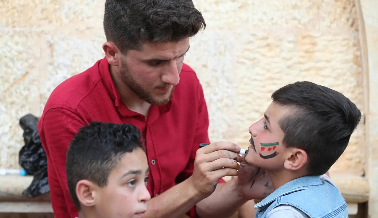 Seorang pria Suriah melukiskan bendera oposisi Suriah di wajah seorang anak selama perayaan Idul Fitri, hari libur Muslim yang dimulai pada akhir bulan puasa Ramadan, di Binnish di provinsi Idlib yang dikuasai pemberontak di barat laut Idlib (26/5/2020). (AFP Photo/Omar Haj Kadour)