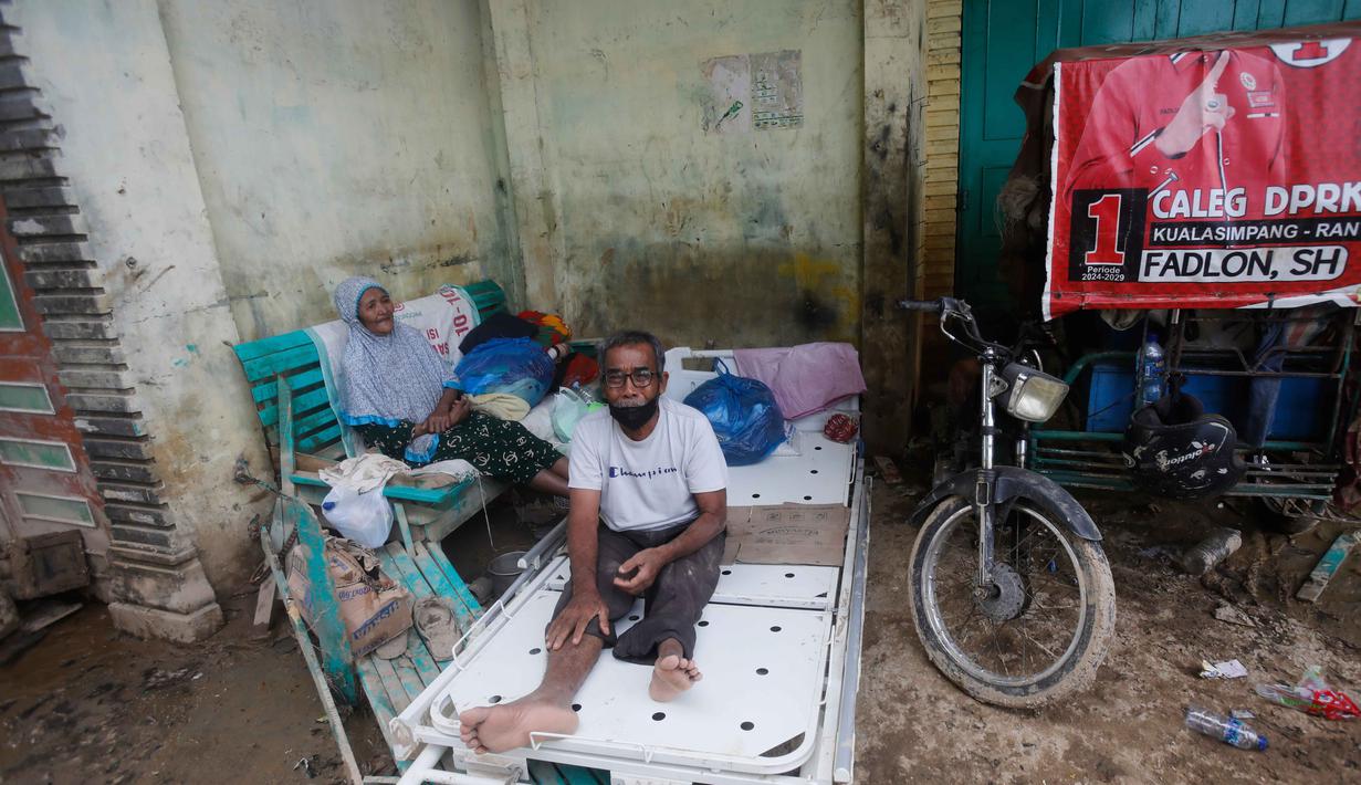 Sejumlah puing-puing, baik itu pohon, sisa material banjir hingga kendaraan yang terguling, masih berserakan di berbagai titik. Kondisi ini menyulitkan akses dan aktivitas warga. Tampak dalam foto, Tengku Rusli, 67 tahun, tengah, bersam istrinya, Tengku Mintan, 64 tahun, yang kehilangan rumah akibat banjir, beristirahat di tempat penampungan sementara mereka di beranda sebuah bangunan di Aceh Tamiang, Pulau Sumatra, Kamis 4 Desember 2025. (AP Photo/Binsar Bakkara)