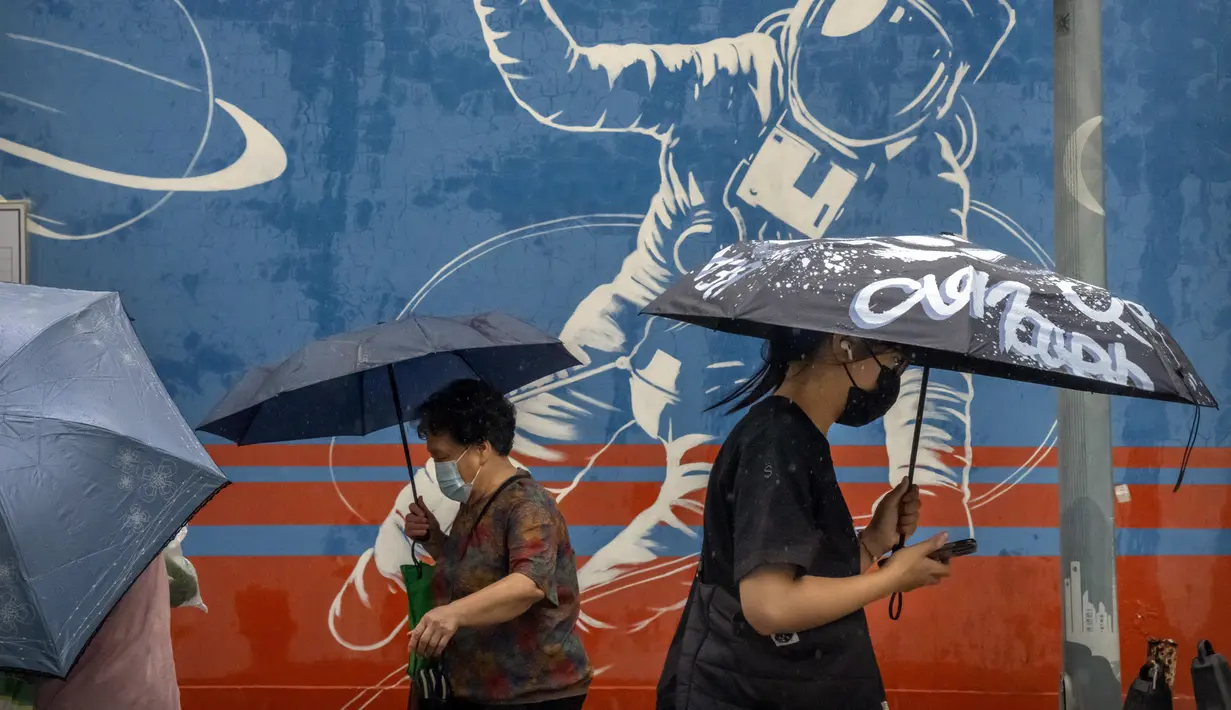 Para komuter yang mengenakan masker dan membawa payung berjalan melewati mural seorang astronot saat hujan di kawasan pusat bisnis di Beijing, China, 12 Juli 2022. Beijing telah membatalkan rencana untuk mengizinkan hanya orang yang divaksinasi yang memasuki ruang publik. (AP Photo/Mark Schiefelbein)