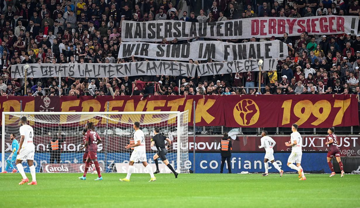 Suporter FC Metz membentangkan spanduk bertulisakan "Pemenang derby Kaiserlautern, kami berada di final" untuk mendukung FC Kaiserlautern yang mencapai final Piala DFB Pokal pada laga lanjutan Liga Prancis 2023/2024 melawan RC Lens di Saint-Symphorien Stadium, Longeville-les-Metz, Prancis, Sabtu (13/04/2024) dini hari WIB. (AFP/Jean-Christophe Verhaegen)