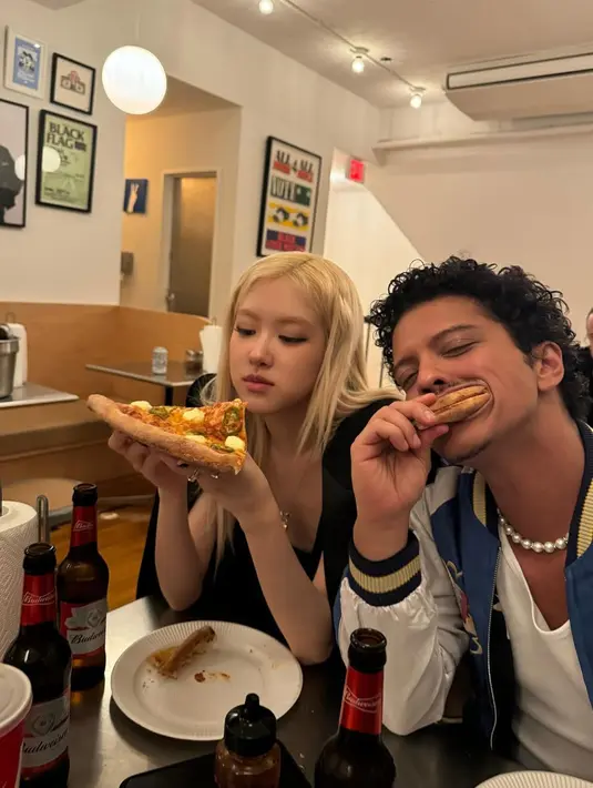 Sementara itu, Bruno Mars tampil dengan gaya playful khasnya. Ia mengenakan jaket varsity biru dengan aksen putih, dipadukan dengan kalung mutiara yang menjadi ciri khas penampilannya yang penuh karakter. Bruno tampak santai dan ceria, menggigit bagian crust pizza sambil tersenyum, memperlihatkan suasana akrab di antara mereka. [@roses_are_rosie].