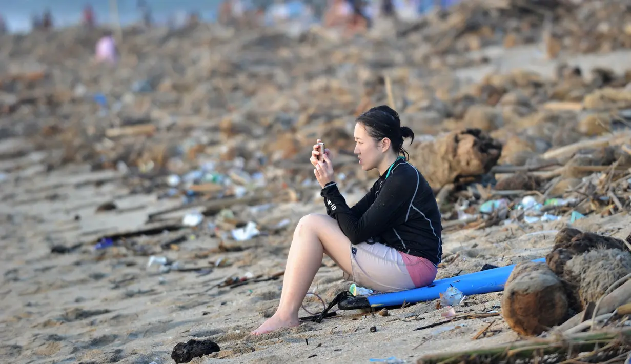 Seorang turis bermain ponsel dekat sampah yang terdampar akibat cuaca buruk di Pantai Kuta, Bali, Jumat (15/2). Hujan deras disertai angin kencang yang melanda Bali berdampak pada arus laut yang terus membawa sampah dari daerah lain. (SONNY TUMBELAKA/AFP)