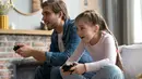 Seorang ayah bersama putrinya sedang bermain video game di dalam rumah, mereka terlihat begitu bersemangat mengalahkan satu sama lain. (OPOLJA/ Shutterstock.com)