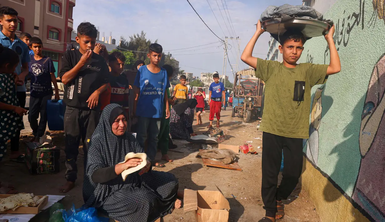 Seorang anak laki-laki Palestina berjalan melewati orang-orang yang sedang membuat roti tidak beragi di lokasi penampungan bagi keluarga pengungsi, Jalur Gaza Selatan, 27 Oktober 2023. (SAID KHATIB/AFP)