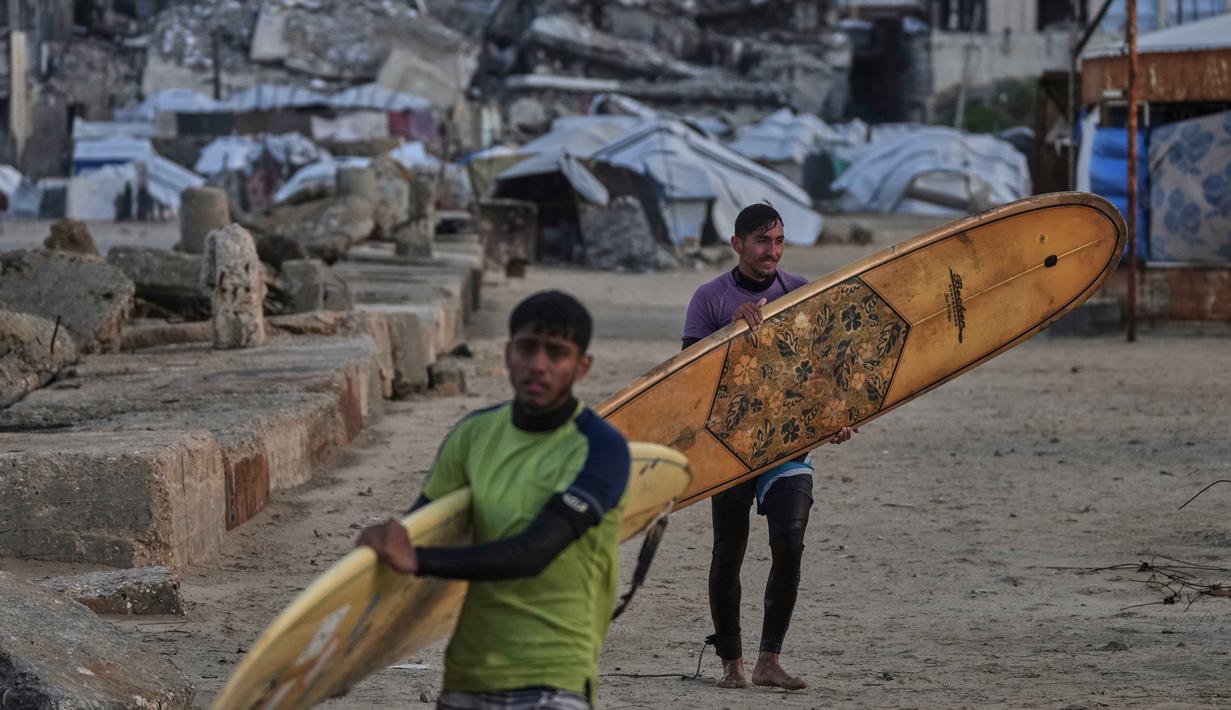 Dan, kawasan pantai di Kota Gaza menjadi salah satu sumber kebahagiaan berharga bagi warga Palestina yang ingin melepaskan diri dari tekanan situasi selama ini. Tampak dalam foto, warga Palestina, Khalil Abu Jayyab (depan) dan Tahseen Abu Assi membawa papan selancar mereka saat memasuki area sepanjang garis pantai di Kota Gaza, Minggu, 28 Desember 2025. (AP Photo/Jehad Alshrafi)