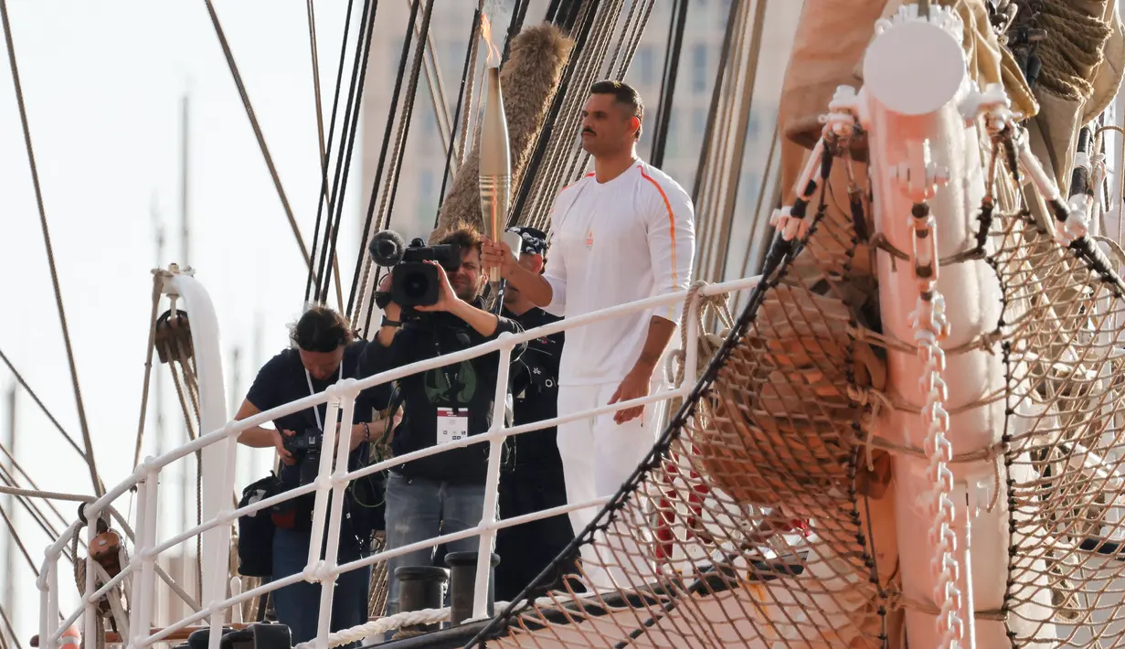 Perenang Prancis, Florent Manaudou memegang obor di atas kapal abad ke-19 bernama Belem saat upacara penyambutan kedatangan api Olimpiade dan Paralimpiade Paris 2024 di Vieux-Port (Pelabuhan Tua) Marseille, Pranci, Rabu (08/05/2024) waktu setempat. (AFP/Pool/Ludovic Marin)