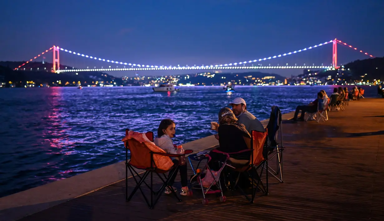 Orang-orang menikmati malam di sebelah pantai Bosphorus ketika jembatan Fatih Sultan Mehmet terlihat di Istanbul (6/7/2020). Jembatan Fatih Sultan Mehmet, yang dikenal dengan sebutan Jembatan Bosporus Kedua, adalah sebuah jembatan di Istanbul, Turki yang melintasi selat Bosporus. (AFP/Ozan Kose)