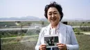 Untuk mendapatkan izin pembangunan kafe ini, pemiliknya harus membangun properti dengan bunker dan posisi yang dibentengi untuk tank. Tampak dalam foto yang diambil pada tanggal 24 April 2025 pendiri kafe Daonsoop, Lee Oh-sook berpose sambil memegang foto kerabatnya yang berkunjung ke Kaesong, Korea Utara pada tahun 2008 sebagai turis. (ANTHONY WALLACE/AFP)