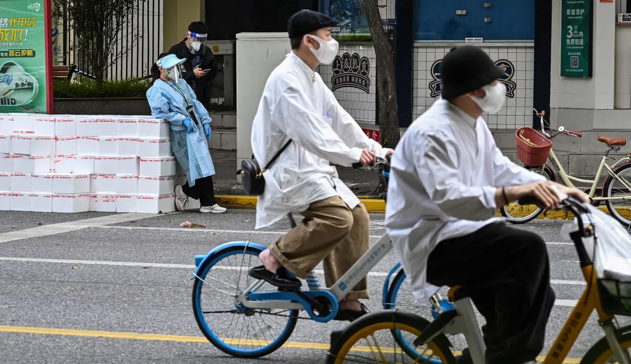 Seorang pekerja yang mengenakan alat pelindung diri berdiri dekat kotak untuk dikirim sementara dua orang mengendarai sepeda saat perberlakuan lockdown karena virus corona COVID-19 di Distrik Jing'an, Shanghai, China, Rabu (18/5/2022). (Hector RETAMAL/AFP)