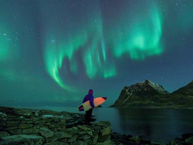 Seorang peselancar menatap Aurora sebelum beraksi di Lofoten Islands, Norwegia, (9/3/2018). Suhu udara minus 13°C dan suhu diatas air sekitar 4°C. (AFP/Oliver Morin)