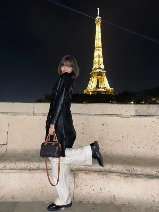 Terlihat berpose dengan latar Menara Eiffel, Lisa BLACKPINK tampil mengenakan set atasan dan bawahan bernuansa putih, yang ditumpuk dengan long coat hitam dengan detail kancing emas. Kali ini, ia membawa small boston in triomphe canvas dan calfskin mini bag dari Celine.