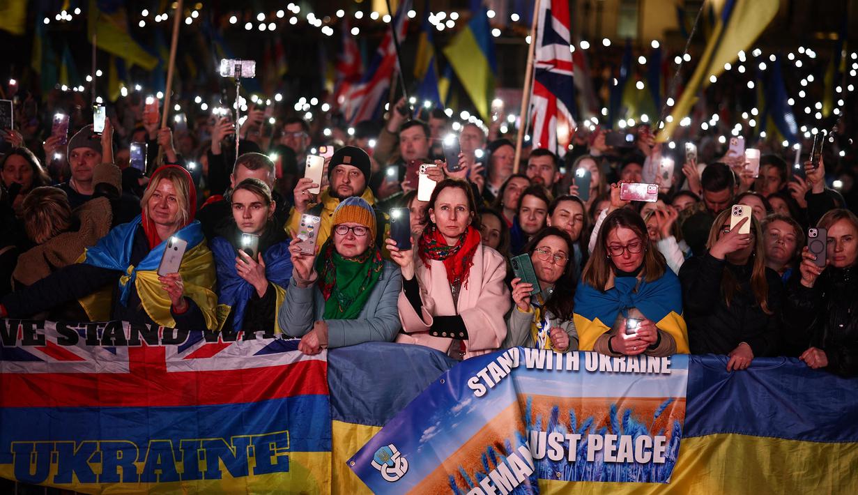 Orang-orang memegang plakat, bendera nasional Ukraina, dan menyalakan lampu ponsel mereka saat berkumpul untuk mendukung Ukraina yang menandai tahun ketiga invasi Rusia di Trafalgar Square, pusat kota London, pada 24 Februari 2025. (HENRY NICHOLLS/AFP)