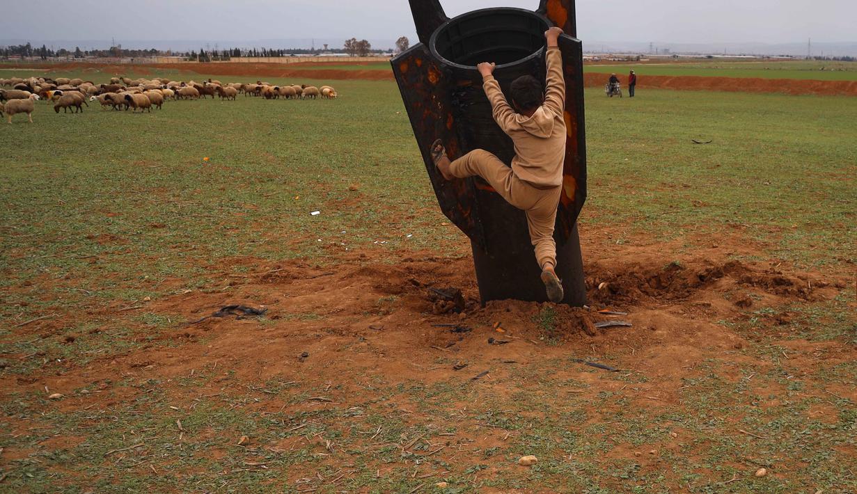 Seorang anak laki-laki mempertaruhkan nyawanya, di tengah bahaya amunisi, dengan mencoba memanjat proyektil Iran yang belum meledak di lapangan terbuka di pinggiran Qamishli, Suriah timur, Rabu 4 Maret 2026. Sebuah proyektil rudal Iran yang belum meledak (unexploded ordnance) mendarat di lapangan terbuka di pinggiran Qamishli, timur laut Suriah. (AP Photo/Baderkhan Ahmad)