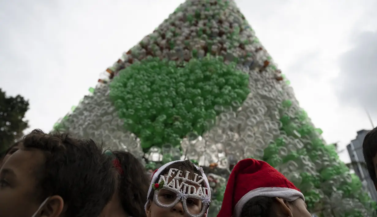 Anak-anak yang mengenakan topi Sinterklas menghadiri upacara penyalaan pohon Natal yang terbuat dari botol plastik daur ulang di Caracas, Venezuela, Selasa (13/12/2022). Pohon Natal yang dibuat dari sekitar 15.000 botol plastik daur ulang ini diinisiasi oleh organisasi nirlaba dan non-pemerintah Oko Spire dan kolaborator. (Yuri CORTEZ / AFP)