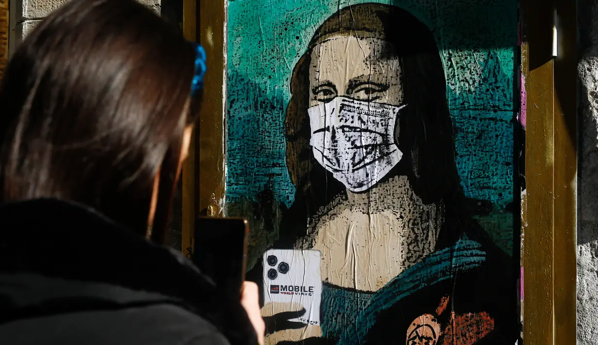 Seorang perempuan mengambil foto poster seniman Italia Salvatore Benintende yang menggambarkan Mona Lisa karya Leonardo da Vinci mengenakan masker dan memegang smartphone yang bertuliskan "Mobile World Virus" di sebuah jalan di kota Barcelona, Spanyol pada Selasa (18/2/2020). (PAU BARRENA/AFP)