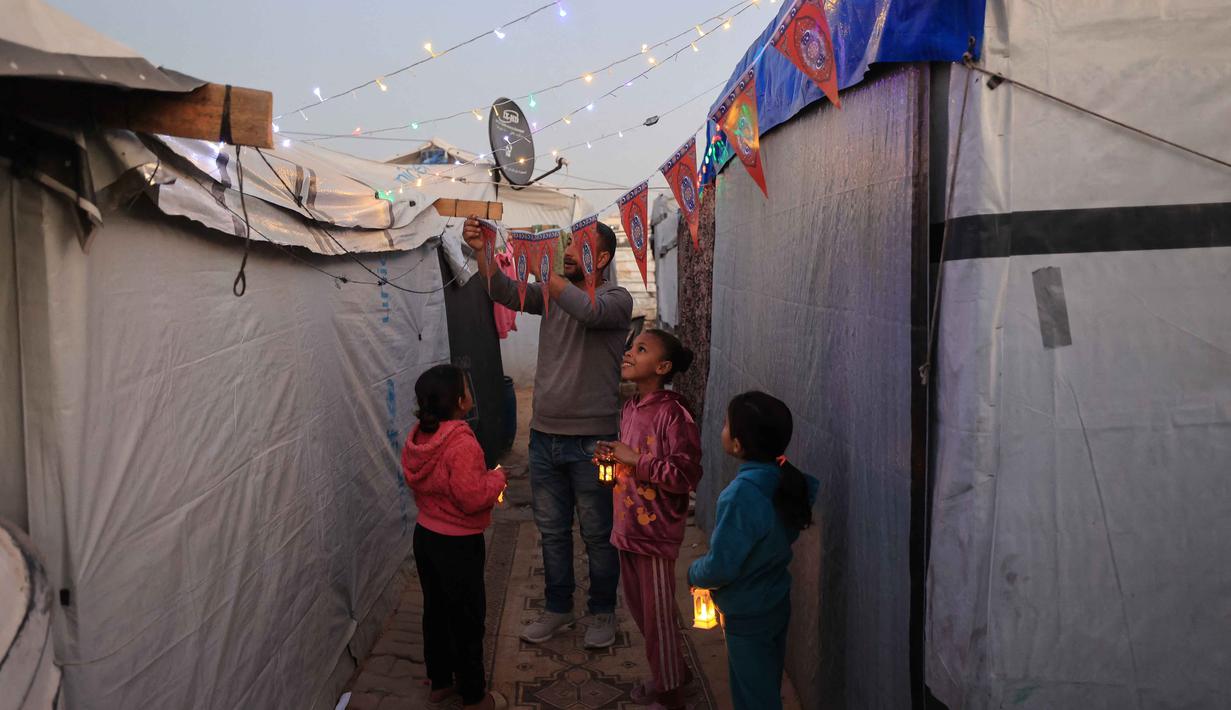 Momen tersebut menjadi ruang kecil untuk merasakan kebersamaan di tengah situasi yang belum sepenuhnya pulih. Tampak dalam foto, Para pengungsi muda Palestina memegang lampion Ramadan saat dekorasi dipasang di samping tenda mereka di kamp pengungsi Bureij di Jalur Gaza tengah pada 15 Februari 2026. (Eyad Baba/AFP)
