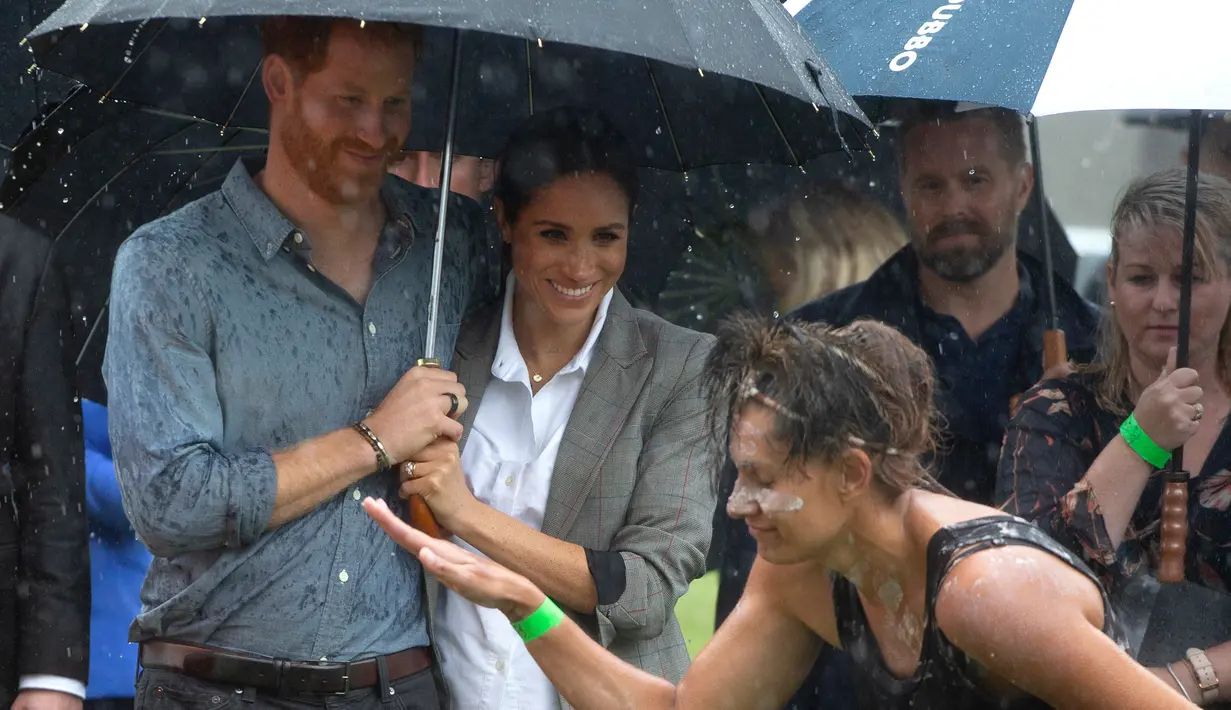 Pangeran Harry dan Meghan Markle berlindung di bawah payung menyaksikan tarian aborigin saat piknik di Victoria Park, Dubbo, Australia, Rabu (17/10). Ini adalah hari kedua Harry dan Markle dalam kunjungannya ke Australia. (Ian Vogler/Pool via AP)