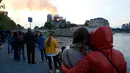Orang-orang menyaksikan api dan asap mengepul dari kebakaran Gereja Katedral Notre-Dame di pusat kota Paris, Prancis, pada Senin (15/4). Api yang melalap bagian atas gereja kuno itu membuat kerumunan baik warga Paris maupun turis tertegun. (AP Photo/Thibault Camus)