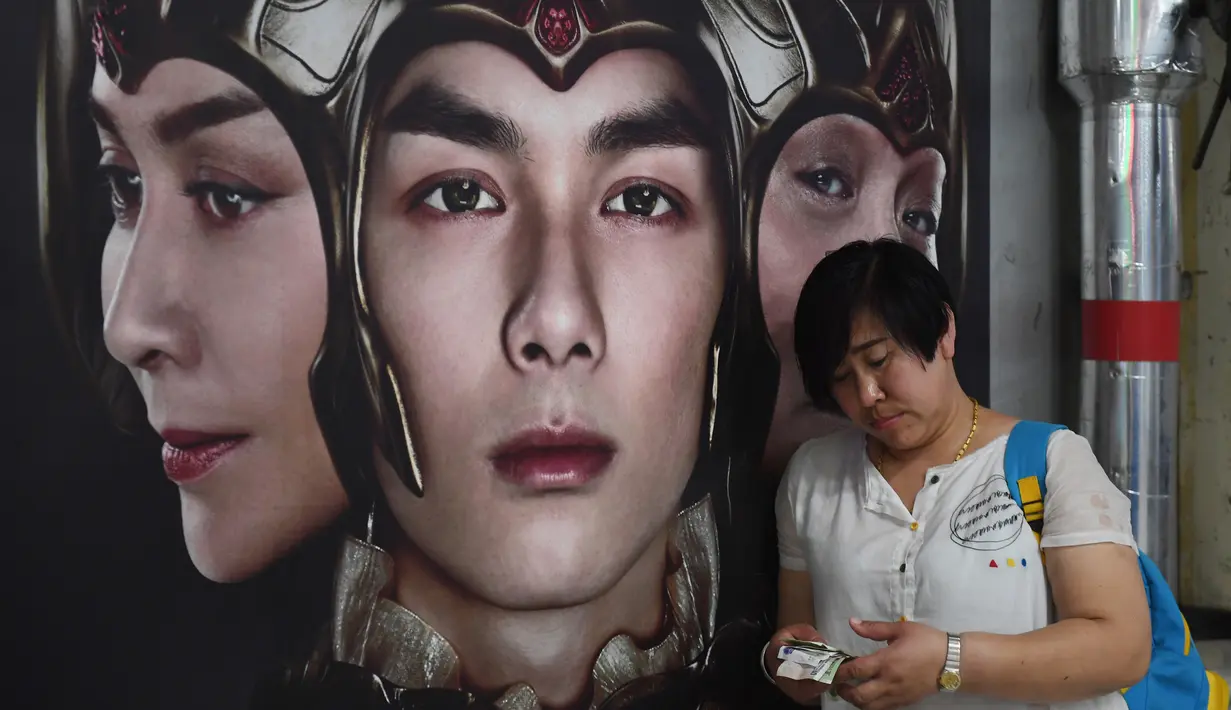Seorang wanita menghitung uang di depan poster film Asura di stasiun kereta bawah tanah di Beijing, China, Selasa (17/7). Asura didasarkan pada mitologi Buddha dan menceritakan kisah dewa-dewa berkepala tiga. (AFP/GREG BAKER)