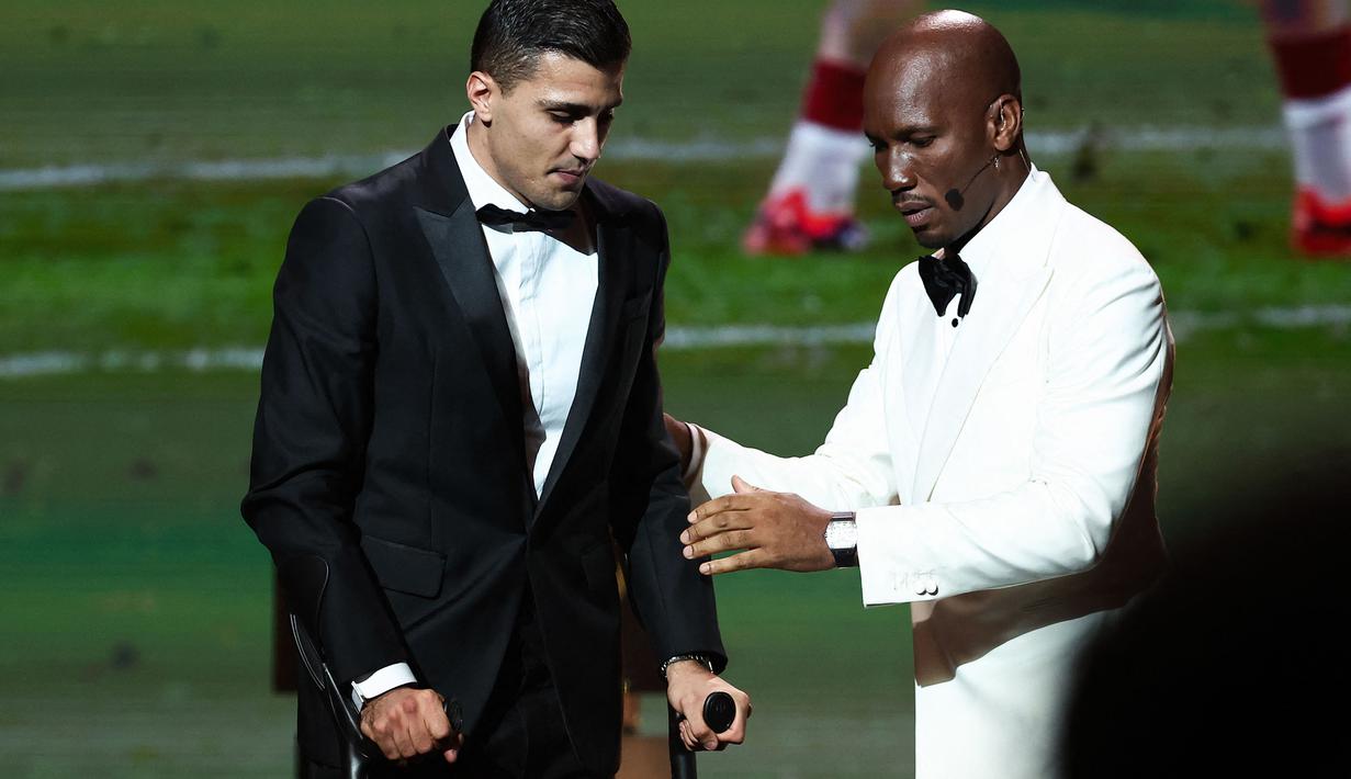 Pemain Manchester City, Rodri (kiri) dipapah oleh mantan pesepak bola, Didier Drogba saat menaiki panggung penghargaan Ballon d'Or 2024 pada acara yang berlangsung di Theatre du Chatelet, Paris, Selasa (29/10/2024) dini hari WIB. (AFP/Franck Fife)