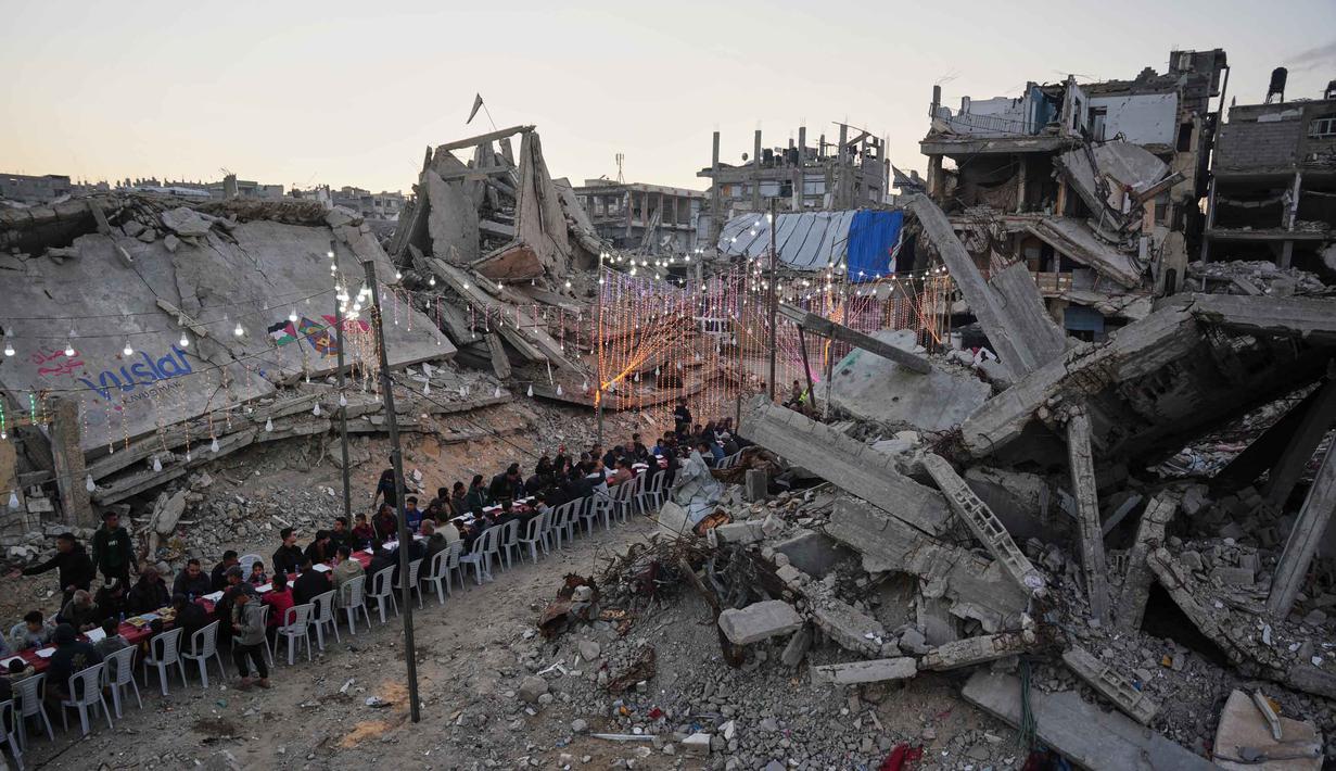 Kelangkaan bahan pokok dan harga yang melambung tinggi membuat banyak warga tidak mampu membeli kebutuhan untuk sahur dan berbuka. Tampak dalam foto, warga Palestina duduk di meja panjang di tengah reruntuhan bangunan yang hancur untuk berbuka puasa bersama, selama bulan suci Ramadan di Khan Younis, Jalur Gaza, Kamis 19 Februari 2026. (AP Photo/Abdel Kareem Hana)