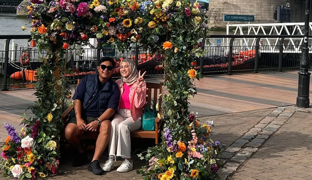 Sebelum masuk dan naik ke Inside Tower Bridge, Nina Zatulini dan sang suami, Chandra Tauphan Ansar berpose dulu di sebuah kursi kayu. Kursi yang berada di area wisata London Bridge itu dikelilingi berbagai macam bunga indah. Pasangan suami istri ini pun terlihat begitu ceria. (Liputan6.com/IG/@ninazatulini22)