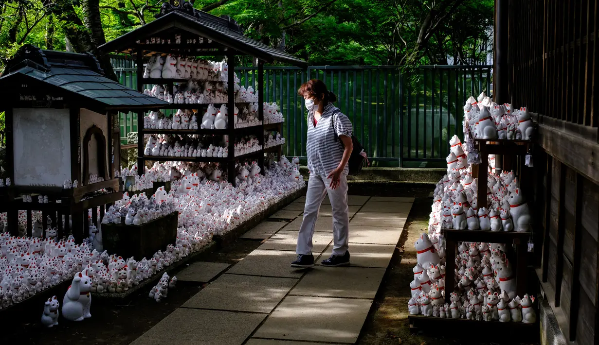 Pengunjung yang mengenakan masker berjalan melewati patung-patung maneki-neko di Kuil Gotokuji, Tokyo, Jepang, Rabu (10/6/2020). Kuil Gotokuji terkenal karena dipercaya sebagai tempat kelahiran maneki-neko, kucing pembawa keberuntungan. (Philip FONG/AFP)