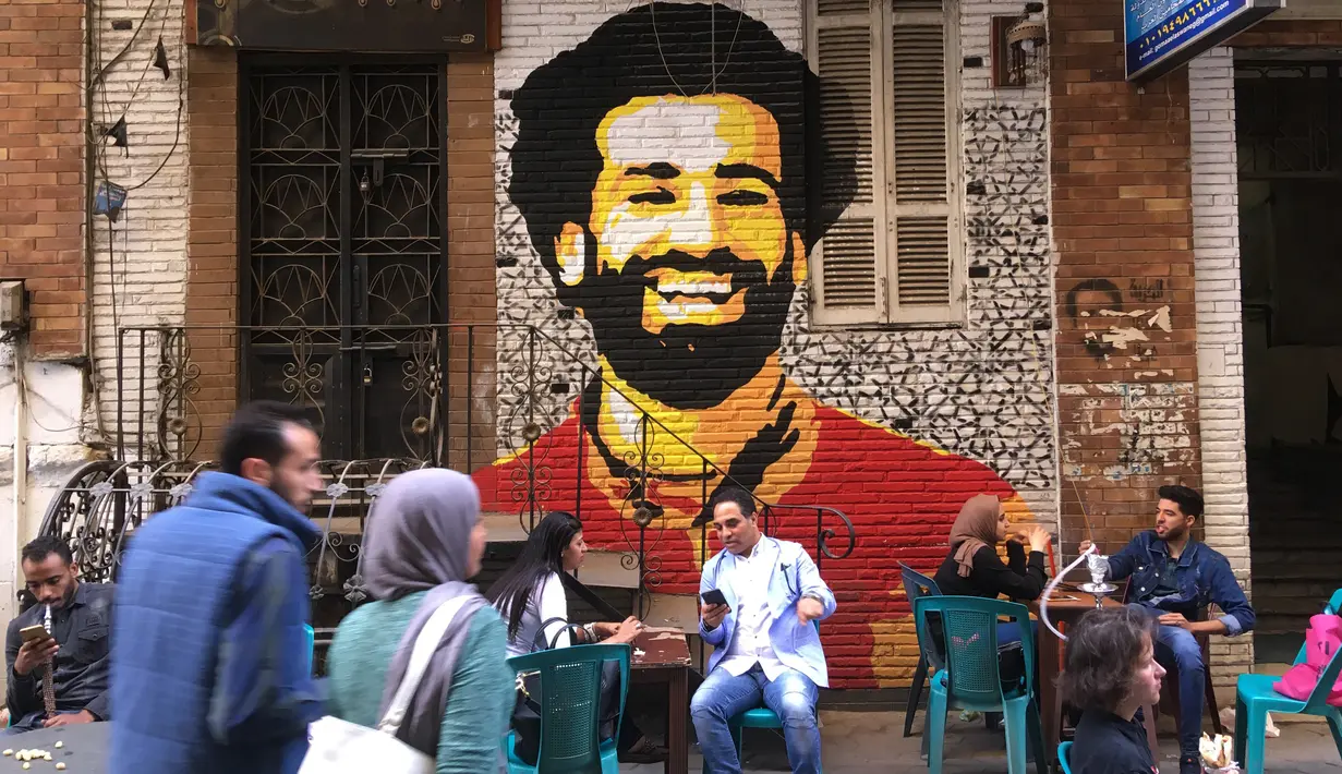 Foto file pada 4 April 2018 menunjukkan orang-orang duduk di sebuah kafe di pusat kota Kairo dengan latar belakang lukisan mural yang menggambarkan striker Liverpool, Mohamed Salah. Federasi Sepak Bola Mesir melaporkan, Jumat (13/11/2020), Mohamed Salah dinyatakan positif Covid-19. (Amir MAKAR/AFP)