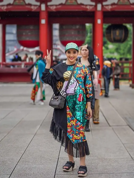 Penampilan colorful dari Ashanty. Ia mengenakan dress dengan motif two-tone yang ramai dan rumbai di ujung dress. Ashanty menambahkan sling bag hitam dan topi hijau yang senada. Foto: Instagram.