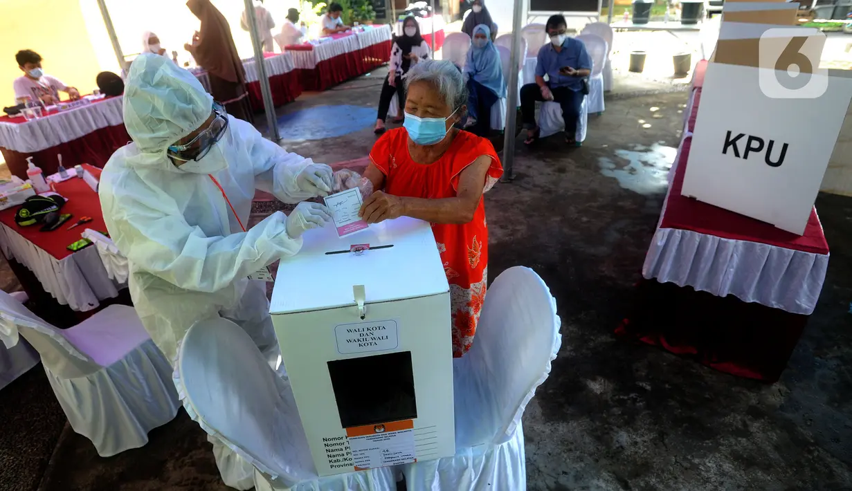 Petugas Kelompok Penyelenggara Pemungutan Suara (KPPS) dengan memakai baju hazmat membantu pemilih memasukkan kertas suara usai menggunakan hak pilihnya dalam Pilkada Tangerang Selatan 2020 di TPS 46, Paku Jaya, Serpong Utara, Rabu (9/12/2020). (merdeka.com/Arie Basuki)