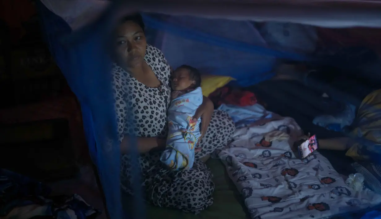 Beberapa kawasan permukiman warga yang kini mengungsi di tenda-tenda darurat dilaporkan masih gelap gulita. Tampak dalam foto, Lia Minarti, 35 tahun, menggendong bayinya, Muhammad Hafid, yang hampir berusia satu bulan, di dalam kelambu di tenda darurat setelah kehilangan rumahnya akibat banjir bandang sekitar tiga minggu sebelumnya, di Aceh Tamiang, pada Selasa 16 Desember 2025. (Yasuyoshi Chiba/AFP)