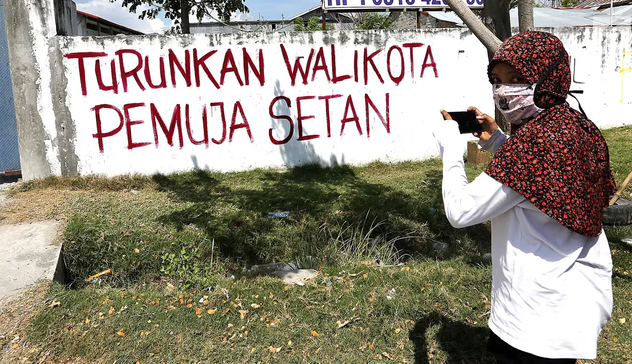 Warga mengabadikan tulisan di dinding dengan pesan Turunkan Walikota Pemuja Setan pasca gempa bumi dan tsunami di Jalan Trans Sulawesi, Palu, Sulawesi Tengah, Kamis (4/10). (Liputan6.com/Fery Pradolo)