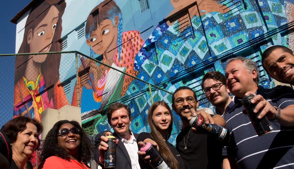 Seniman Brasil, Luna Buschinelli berpose dengan latar belakang mural berjudul 'Contos' di dinding gedung sekolah Rio de Janeiro, (19/6). Mural itu mengisahkan seorang ibu buta huruf yang sedang membacakan buku cerita ke anak-anaknya. (AP/Silvia Izquierdo)
