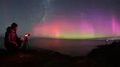 Seorang pria memotret Aurora Australis saat bersinar di cakrawala di atas perairan Danau Ellesmere di pinggiran Christchurch, Selandia Baru pada 24 April 2023. (AFP/Sanka Vidanagama)