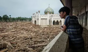 Di antara tumpukan pohon tersebut, terdapat bangunan masjid dengan kubah berwarna hijau yang masih kokoh berdiri. Tampak dalam foto, warga menyaksikan tumpukan pohon tumbang yang tersapu banjir bandang sekitar tiga minggu lalu dari koridor lantai dua yang tergenang air di Pesantren Darul Mukhlisin dan masjid yang terhubung dengan pesantren tersebut di Aceh Tamiang, pada Minggu 14 Desember 2025. (Yasuyoshi CHIBA/AFP)